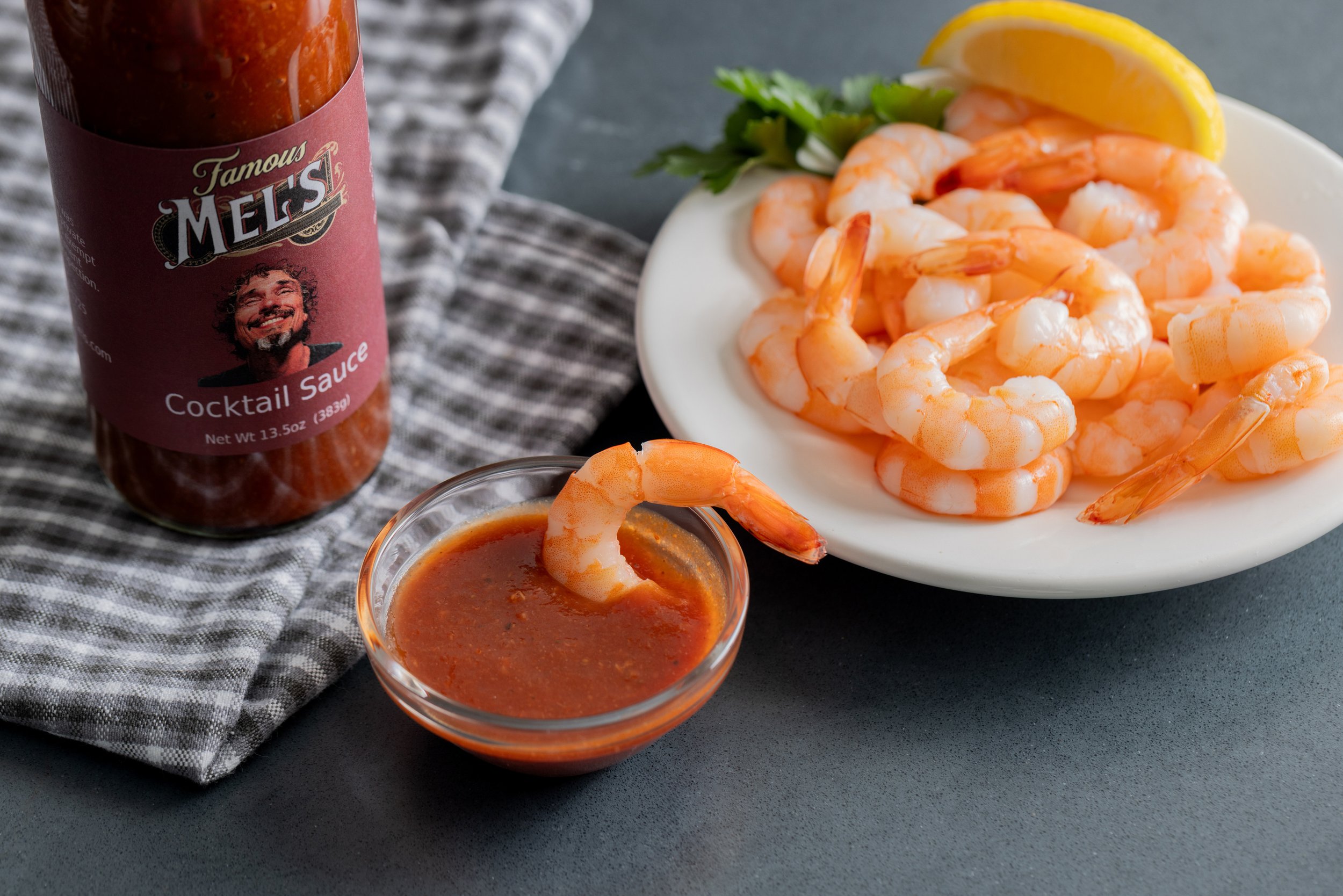 A plate of cooked shrimp with lemon wedges and parsley, a small bowl of cocktail sauce with a shrimp, a bottle of Mel's cocktail sauce, on a checkered cloth.