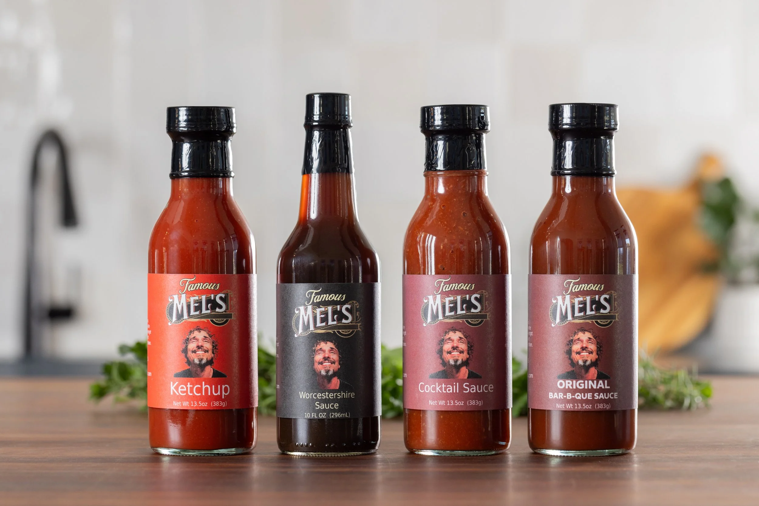 Four bottles of Mel's sauce flavors: ketchup, Worcestershire sauce, cocktail sauce, and original barbecue sauce, arranged on a wooden surface.
