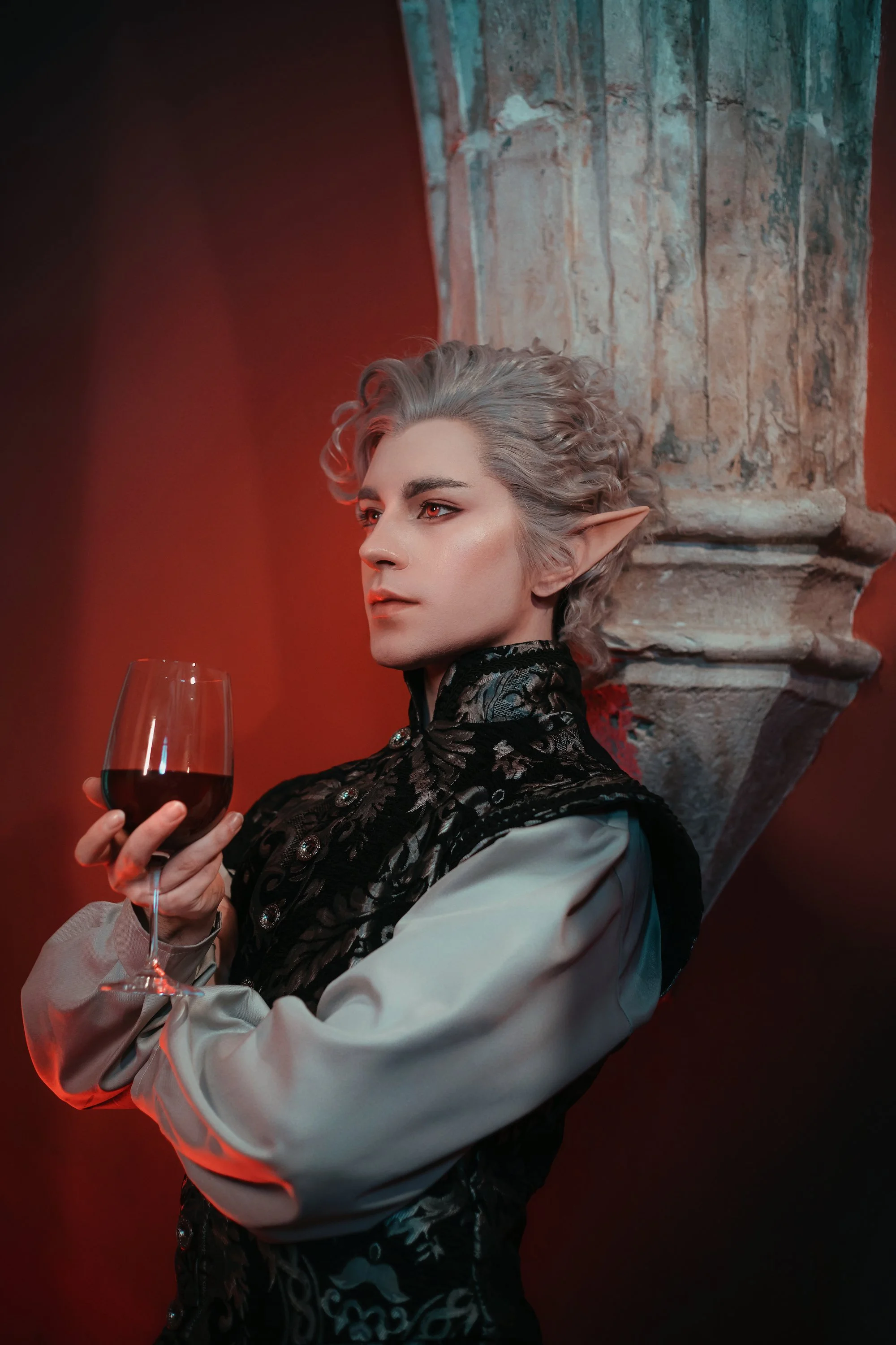 A person with elf-like ears and gray curly hair, dressed in a black embroidered vest and beige shirt, holding a glass of red wine, standing against a red-lit background with a stone column.