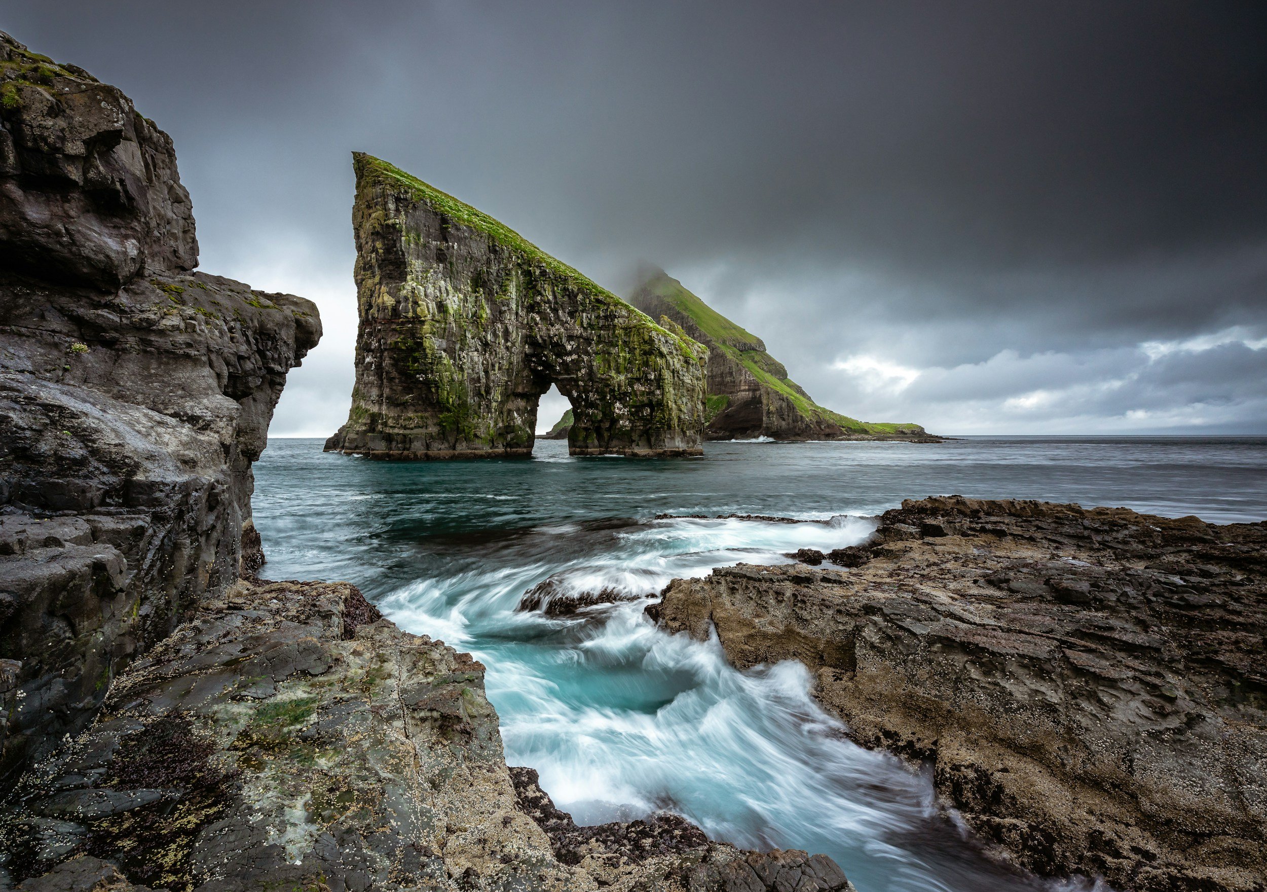 A coastal scene with large rocky formations and a natural rock arch in the ocean, under a cloudy sky.