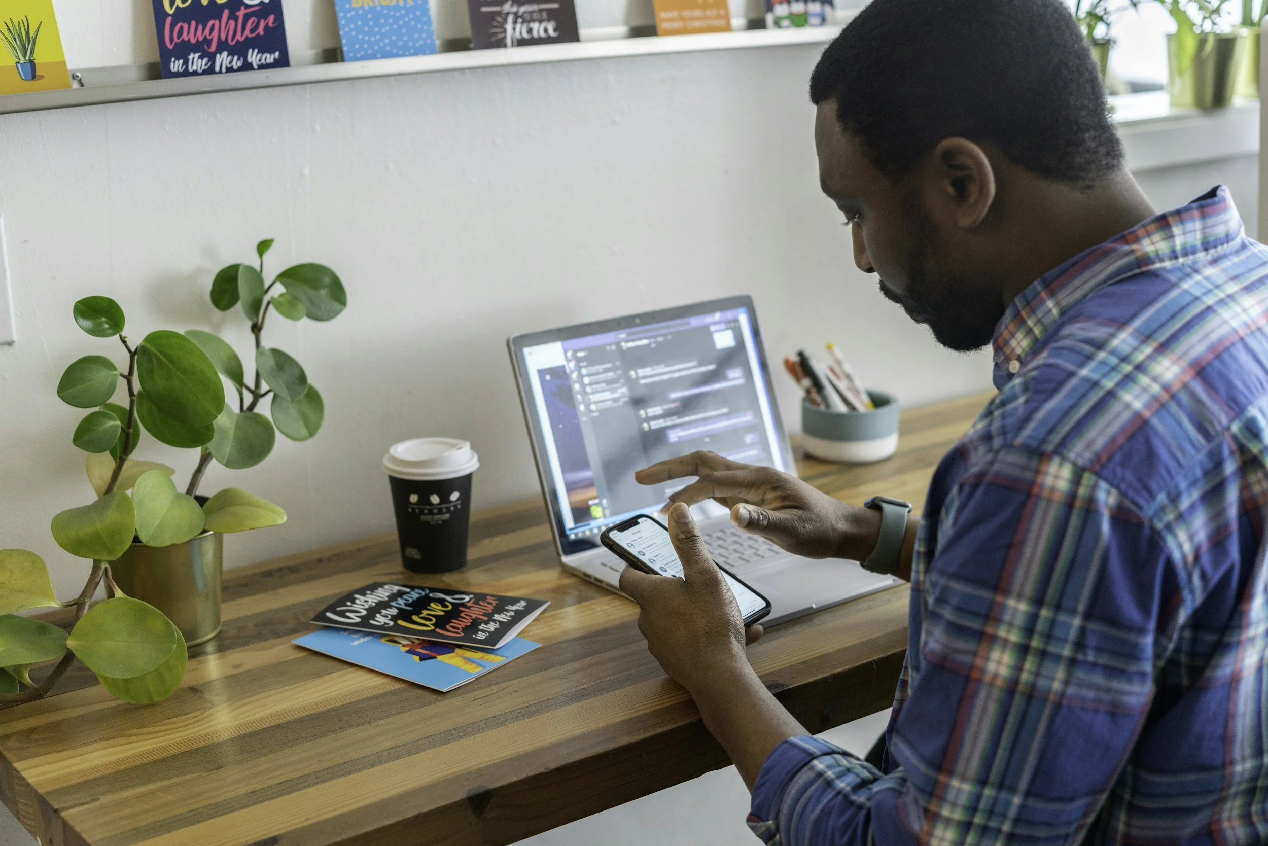A man in a plaid shirt works on a laptop and phone at a wooden desk, with colorful cards, a plant, a coffee cup, and books in the background.
