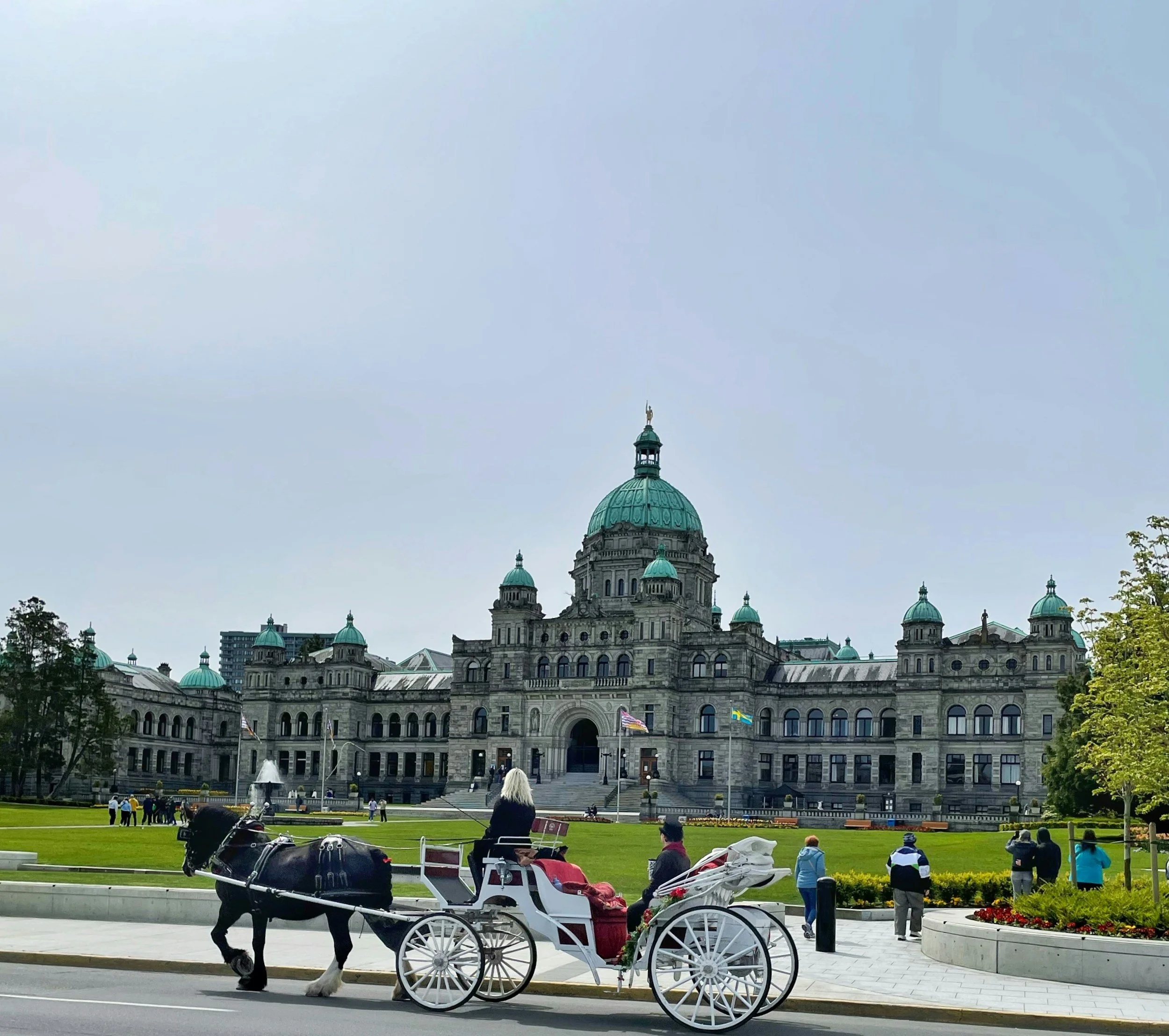 A horse-drawn carriage in front of a historic government building with a prominent dome and multiple smaller domes, populated with people walking and sitting on a grassy area.