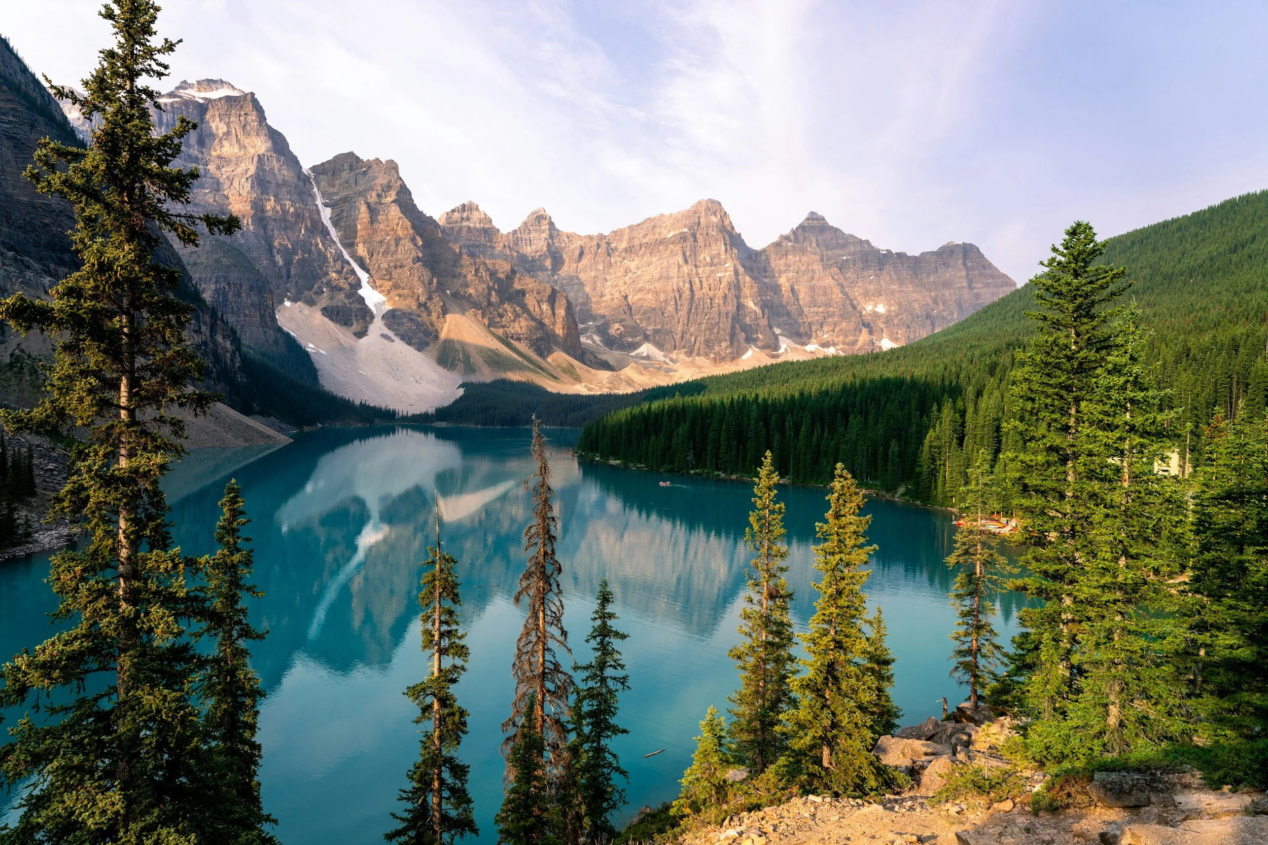 A mountain landscape featuring a large lake with turquoise water, forested hills, and rugged mountains with patches of snow.