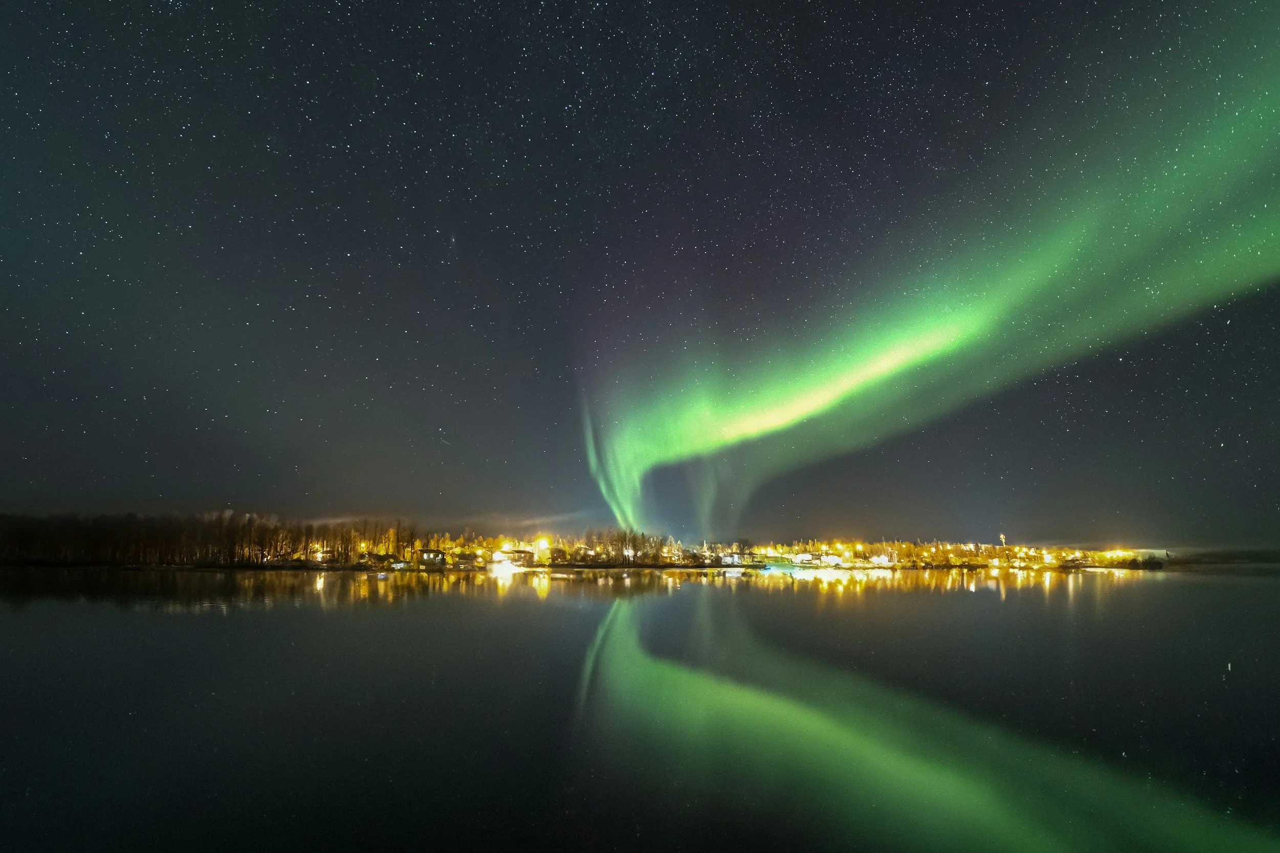 Night sky with stars and green Northern Lights over a small town by a body of water.