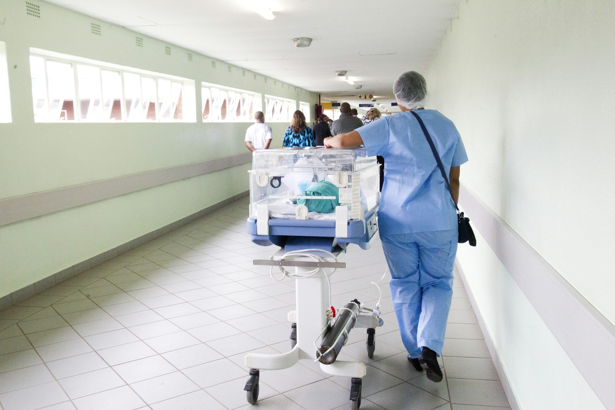 Hospital corridor with medical staff walking, including a nurse pushing a baby incubator, and patients in the background.