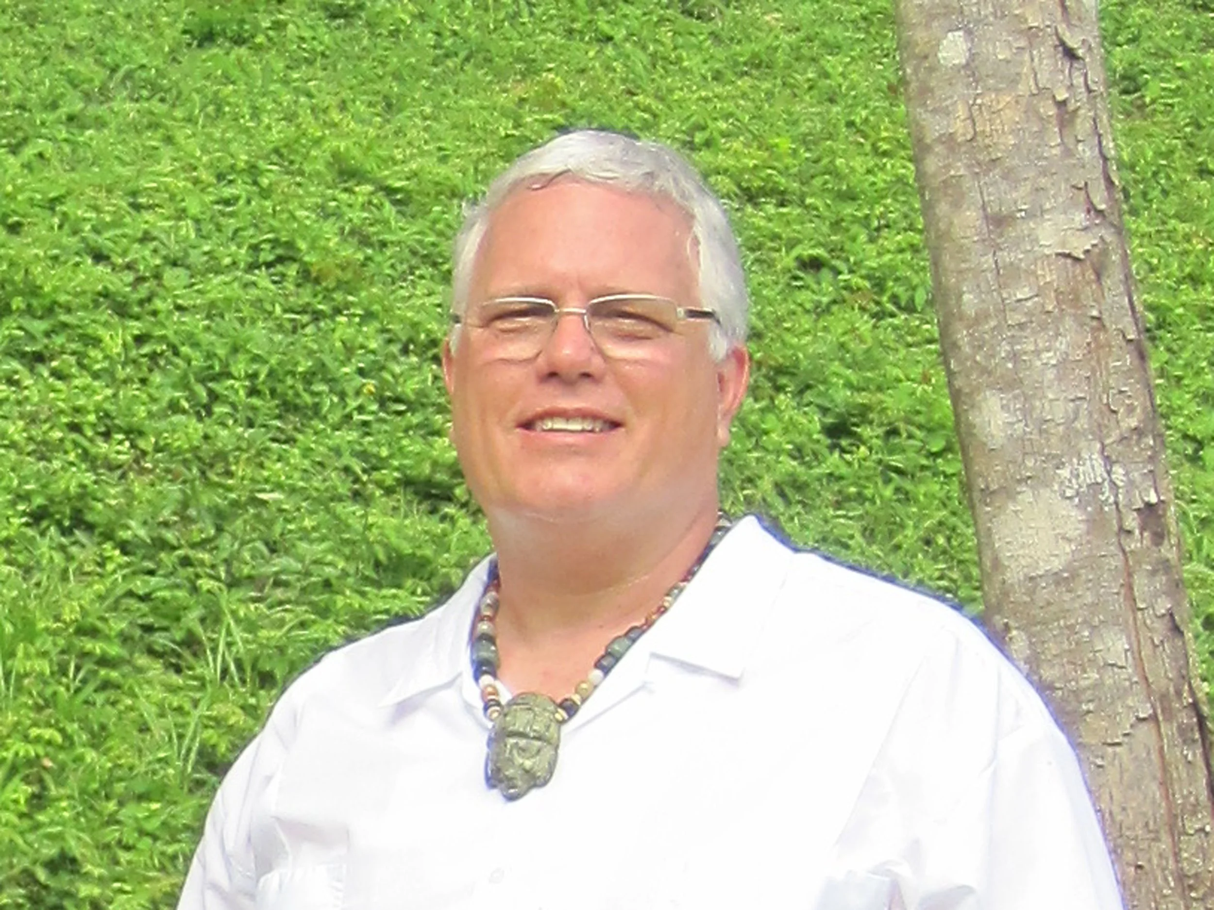 A smiling man with glasses and gray hair standing outdoors near a tree with green foliage in the background.