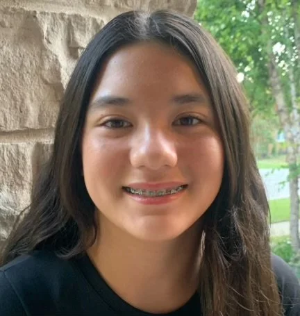 A girl with long dark hair smiling, wearing braces, standing outdoors near a stone wall with trees in the background.