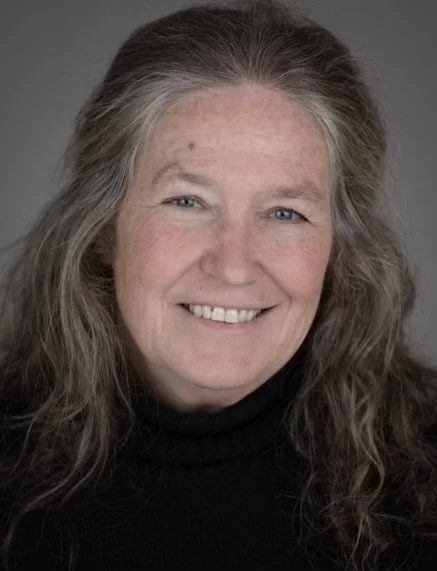 A woman with long gray hair smiling at the camera, wearing a black turtleneck, against a gray background.