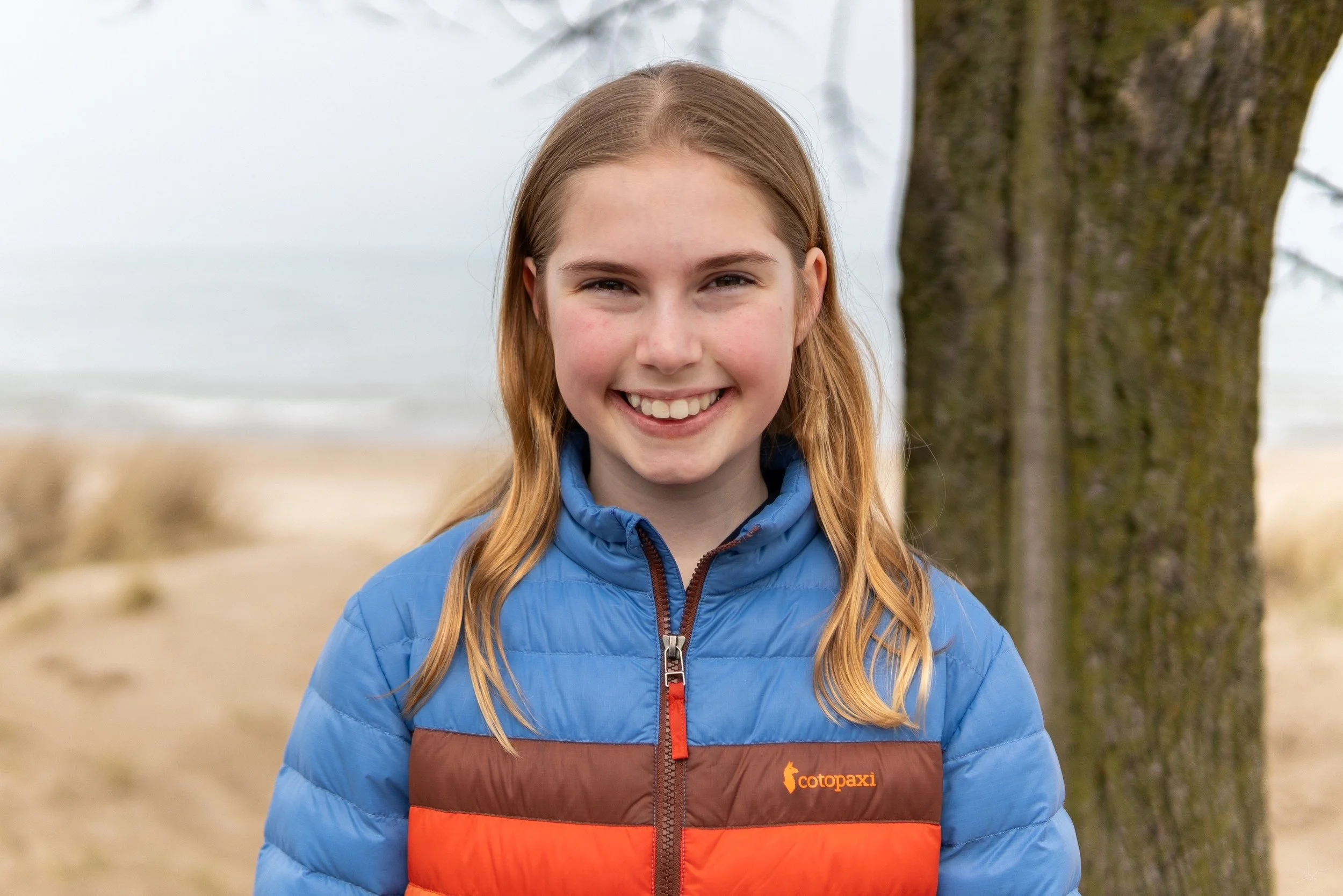 A young girl with long blonde hair smiling outdoors, wearing a blue and orange Cotopaxi jacket, standing next to a tree with a blurred beach and ocean in the background.