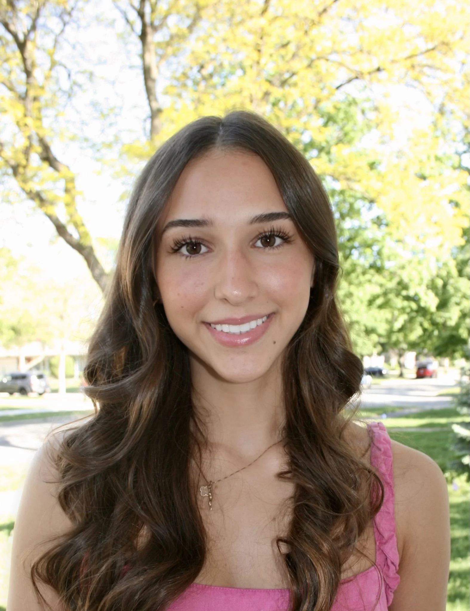 A young woman with long wavy brown hair, smiling, outdoors in daytime with trees and a neighborhood street in the background.