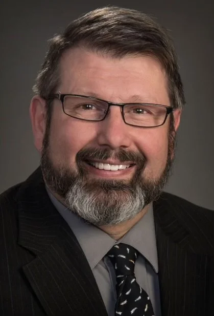 A smiling man with glasses, a beard, and short brown hair, dressed in a dark suit, gray shirt, and patterned tie, against a dark background.