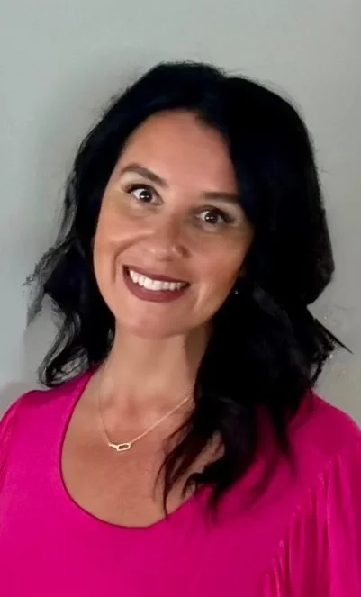 A woman with shoulder-length dark hair, smiling, wearing a pink top and a gold necklace, standing against a light gray background.