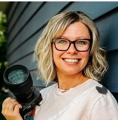 A woman with blonde, shoulder-length hair and glasses smiling while holding a camera with a large lens outdoors.