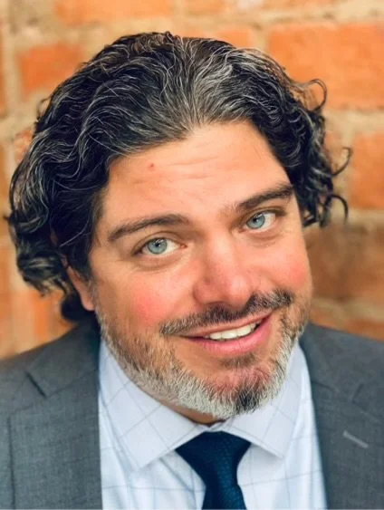 A man with curly dark hair and a beard, smiling, wearing a suit and tie, with a brick wall background.