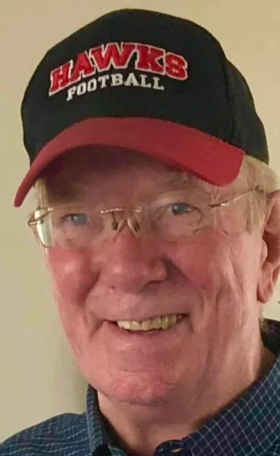 A man wearing glasses and a black and red Hawks Football cap, smiling.