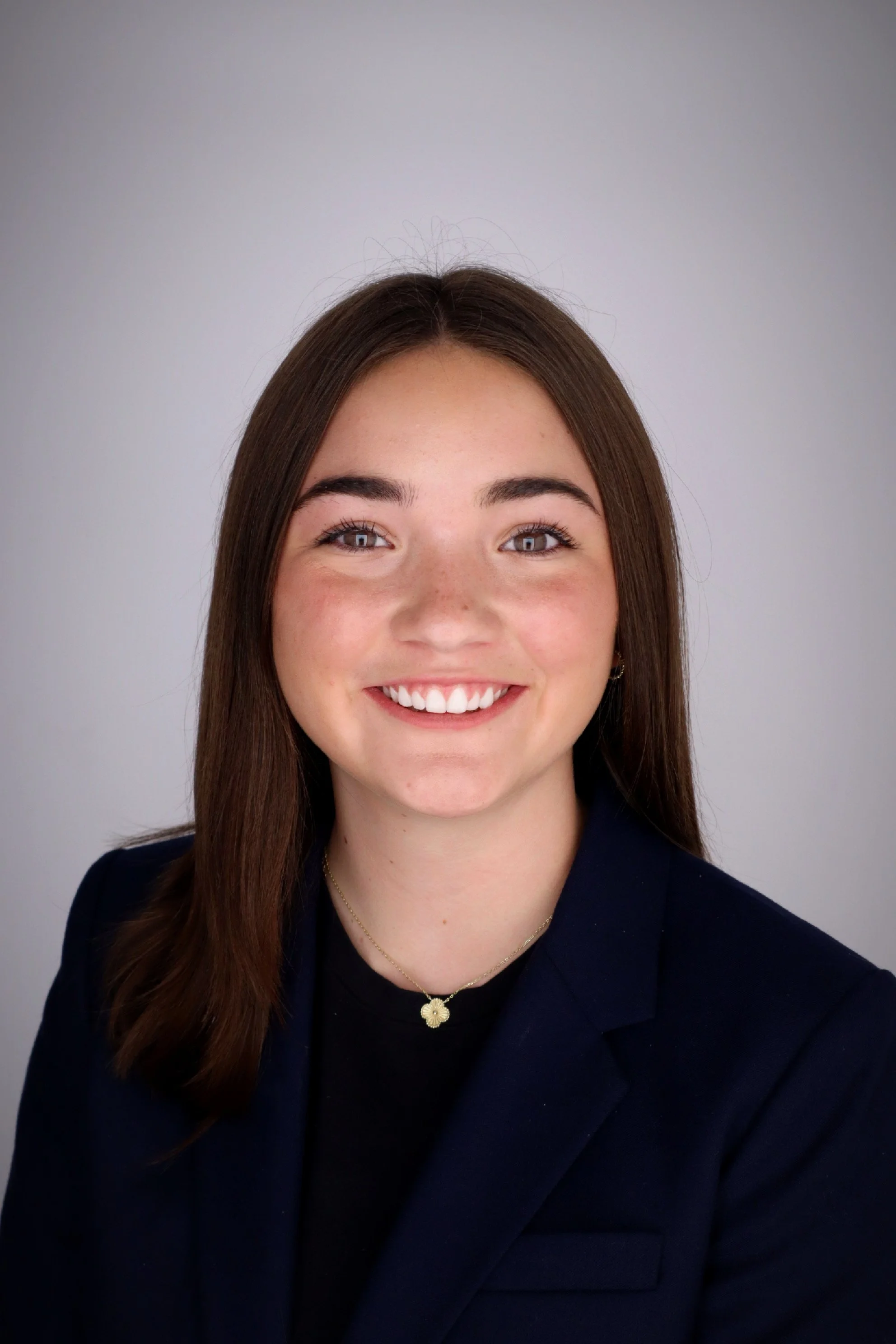 A young woman with long brown hair, wearing a dark blazer and a gold necklace, smiling at the camera against a light gray background.
