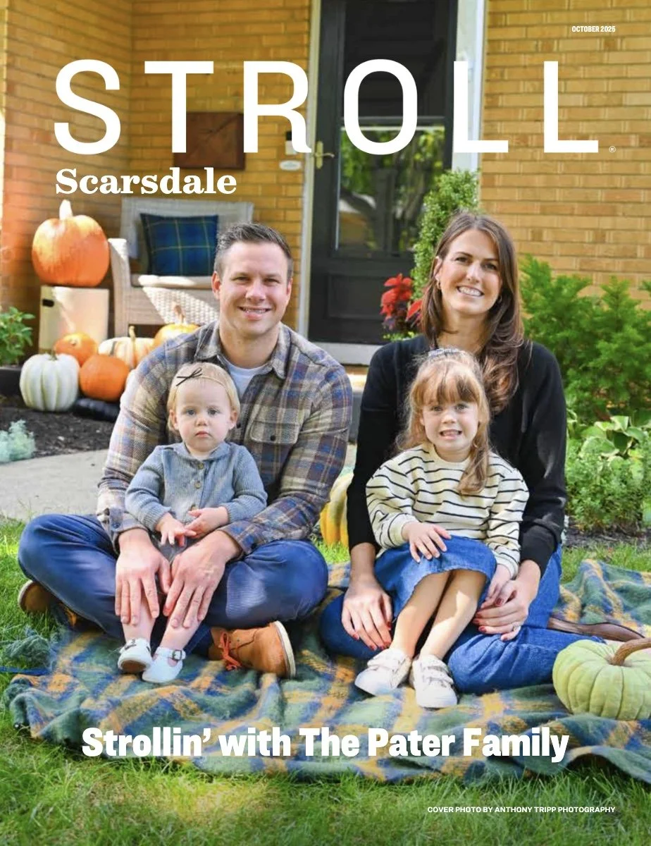 A family of four sitting on a blanket outdoors in front of a house decorated for fall, with pumpkins in the background. The family members include a man, a woman, and two young girls. The man and woman are smiling, while the two girls are seated on the blanket with one smiling and the other with a neutral expression.