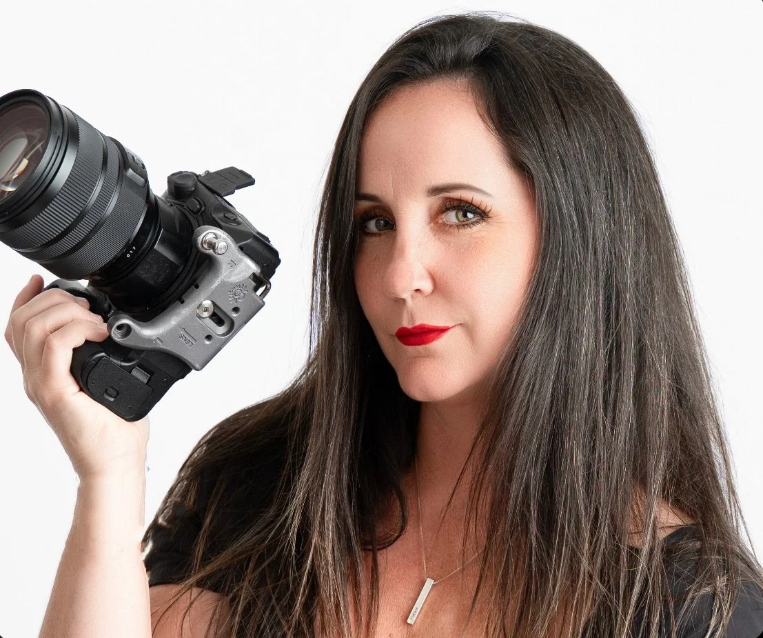 A woman with long dark hair holding a professional camera, wearing red lipstick and a silver necklace, against a plain white background.