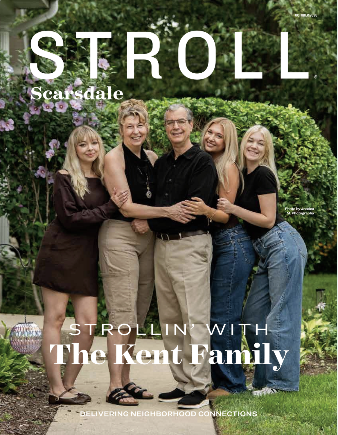 Family portrait of five members standing outdoors in a garden with greenery and flowers, smiling and embracing.