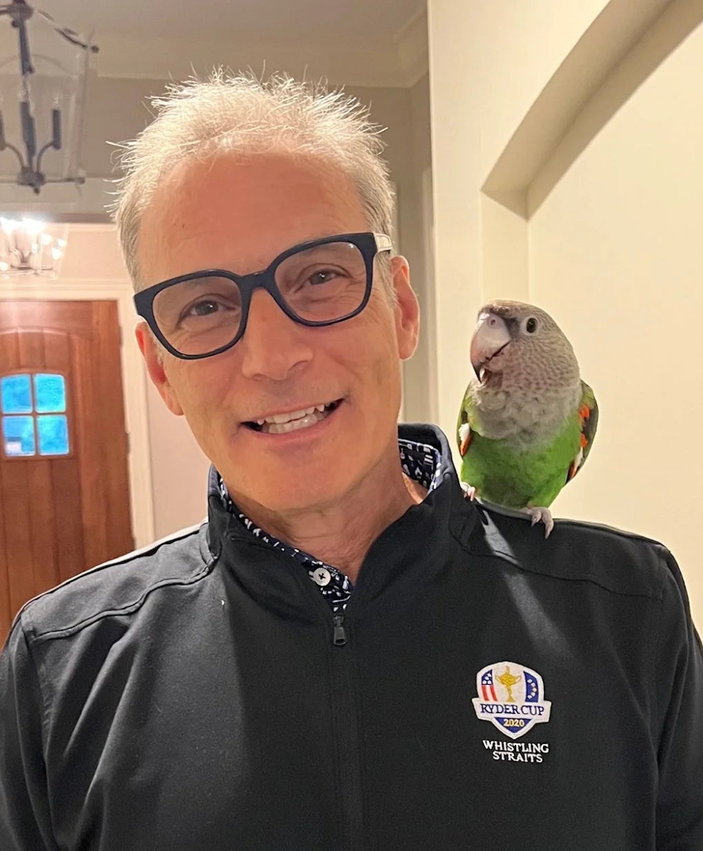 A smiling man wearing black glasses and a black jacket with a Ryder Cup 2020 logo, with a parrot perched on his shoulder in an indoor setting.