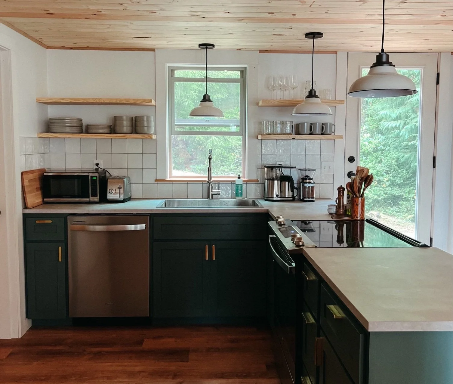 Kitchen with dark green cabinets, wooden countertops, open shelves, window above sink, gray mugs, glasses, and plates, small appliances, and pendant lights.