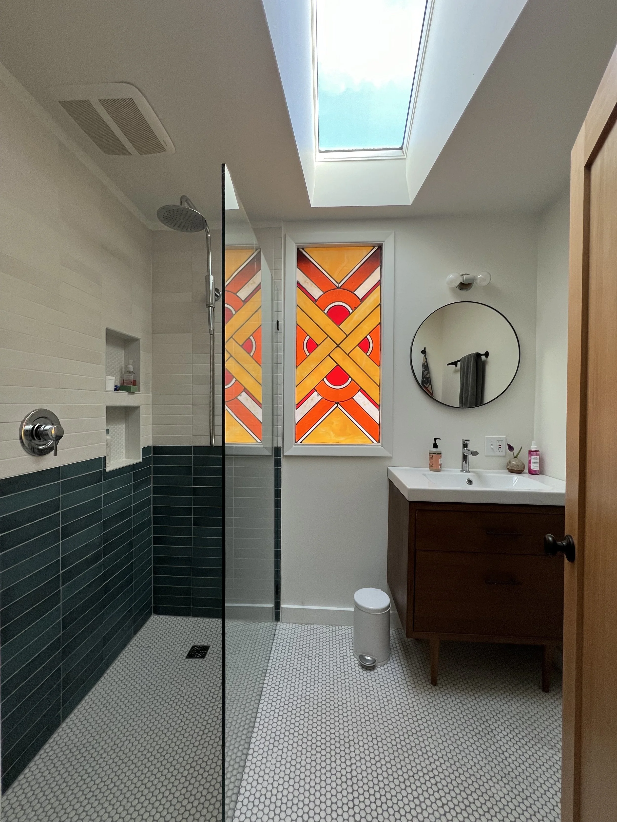 A modern bathroom with a glass shower, a skylight ceiling, a stained glass window with geometric patterns, a round mirror above a wooden vanity, a small trash can, and various toiletries on the sink.