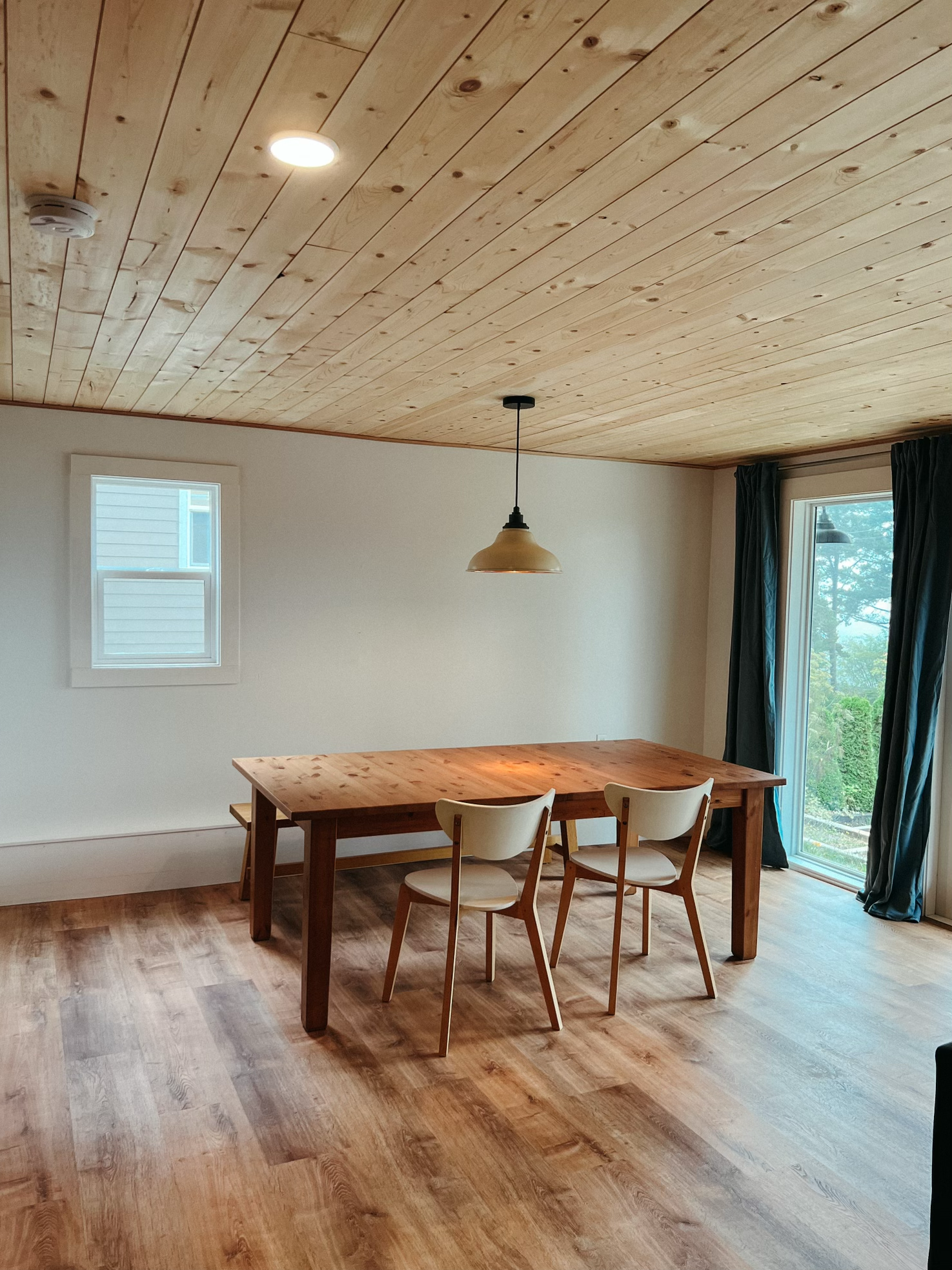 Empty dining room with wooden ceiling, wooden table, two white chairs, a window, a sliding glass door with dark curtains, and a pendant light.