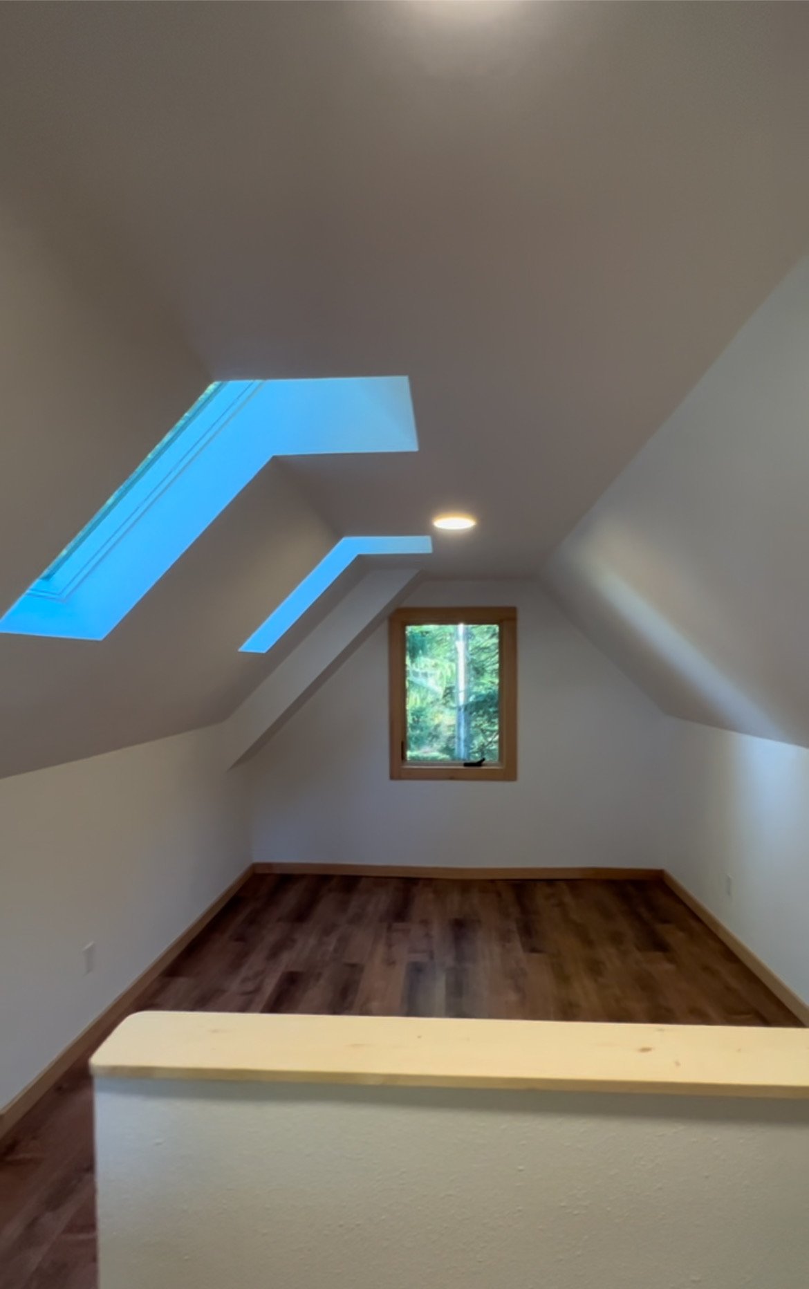 Empty attic room with sloped ceilings, two skylights, a window, hardwood floors, and a small wall