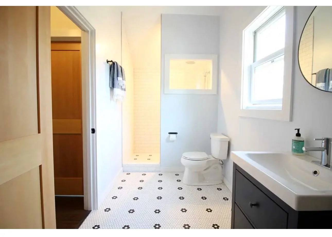 A modern bathroom with white walls, a small window, a toilet, a dark vanity with a white sink, a mirror, and a soap dispenser. There is a walk-in shower with white subway tiles and two towels hanging on the wall. The floor has white tiles with black 