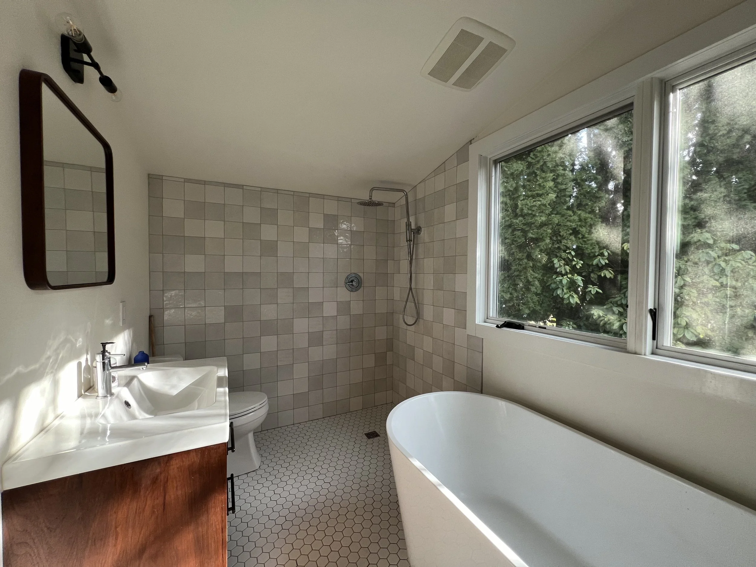 Bathroom with a large window, a bathtub, a floating vanity with a sink, a mirror, a shower area with a tiled wall and rainfall showerhead, and indoor greenery outside the window.
