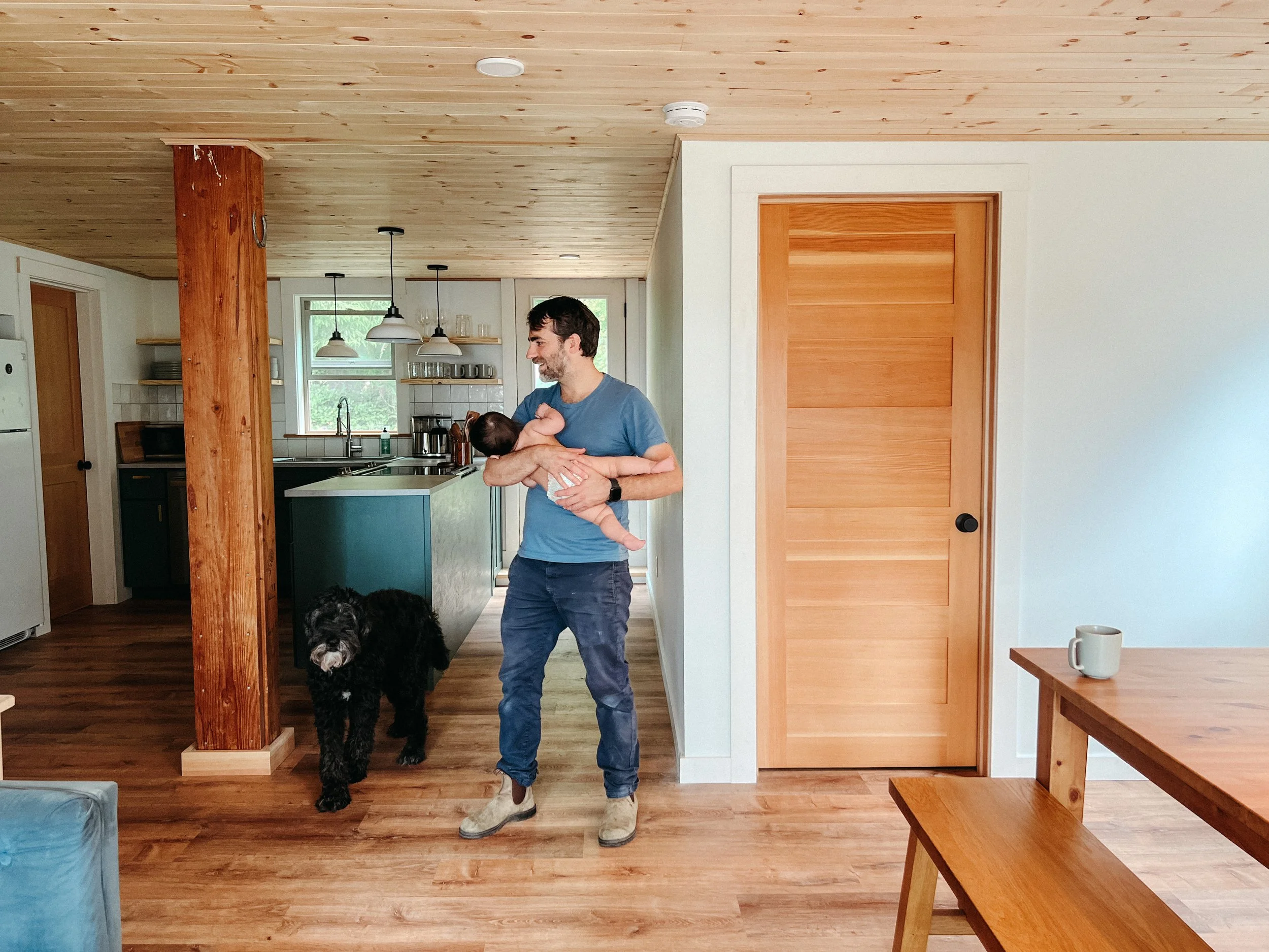 A man holding a baby and a large black dog in a cozy kitchen with wood accents and modern decor.