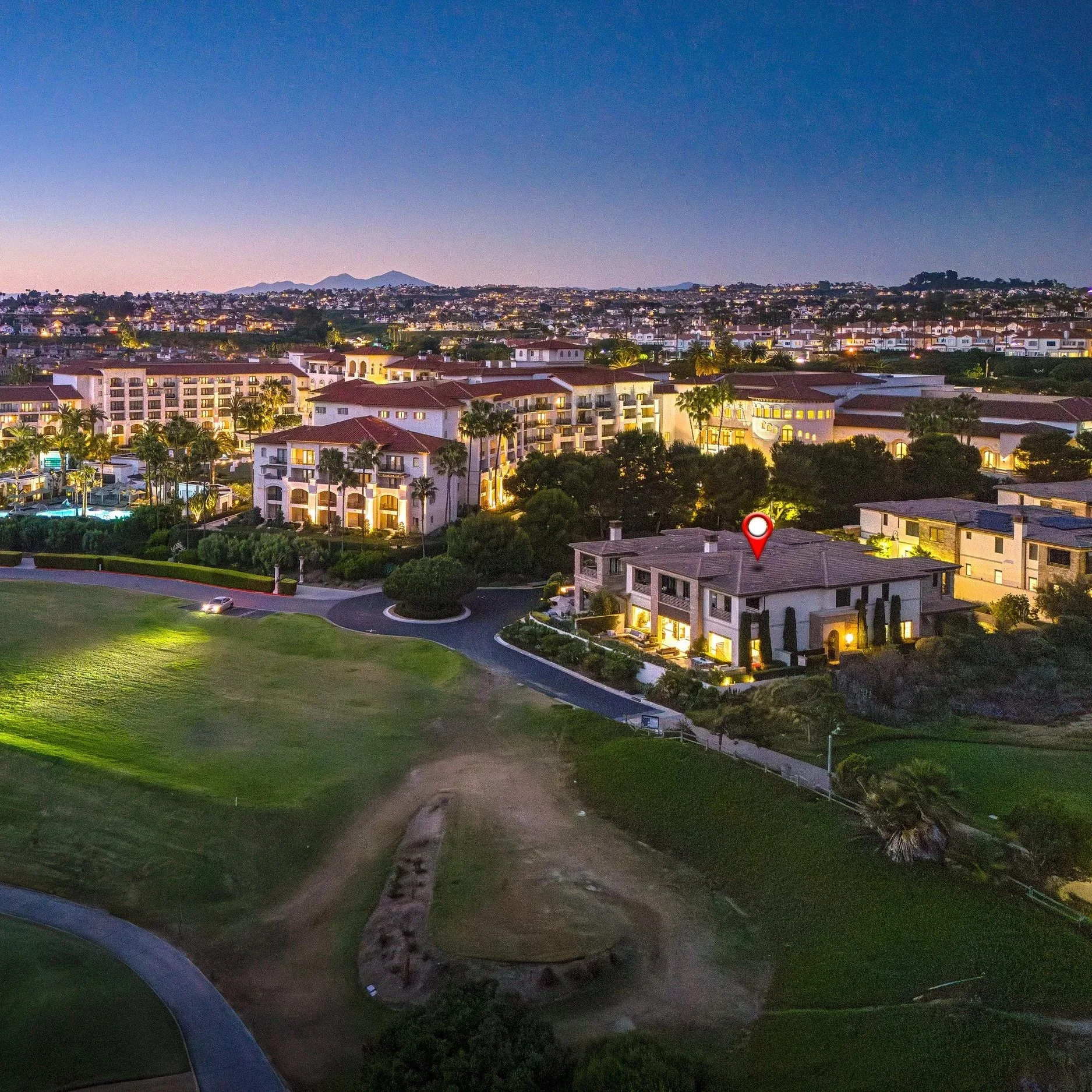 Aerial view of a residential area and golf course at dusk with illuminated buildings and homes, mountains in the background, and a house marked with a red location pin.