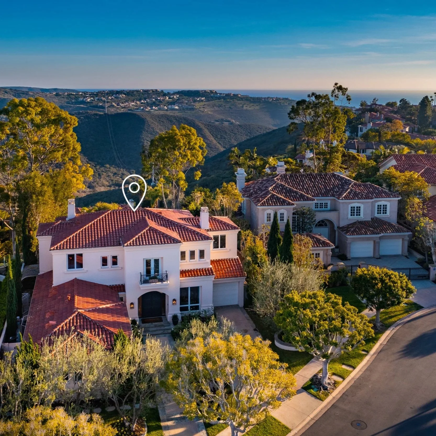 Aerial view of a luxurious house with a red tile roof, surrounded by green trees, overlooking hills with other houses and the ocean in the background.