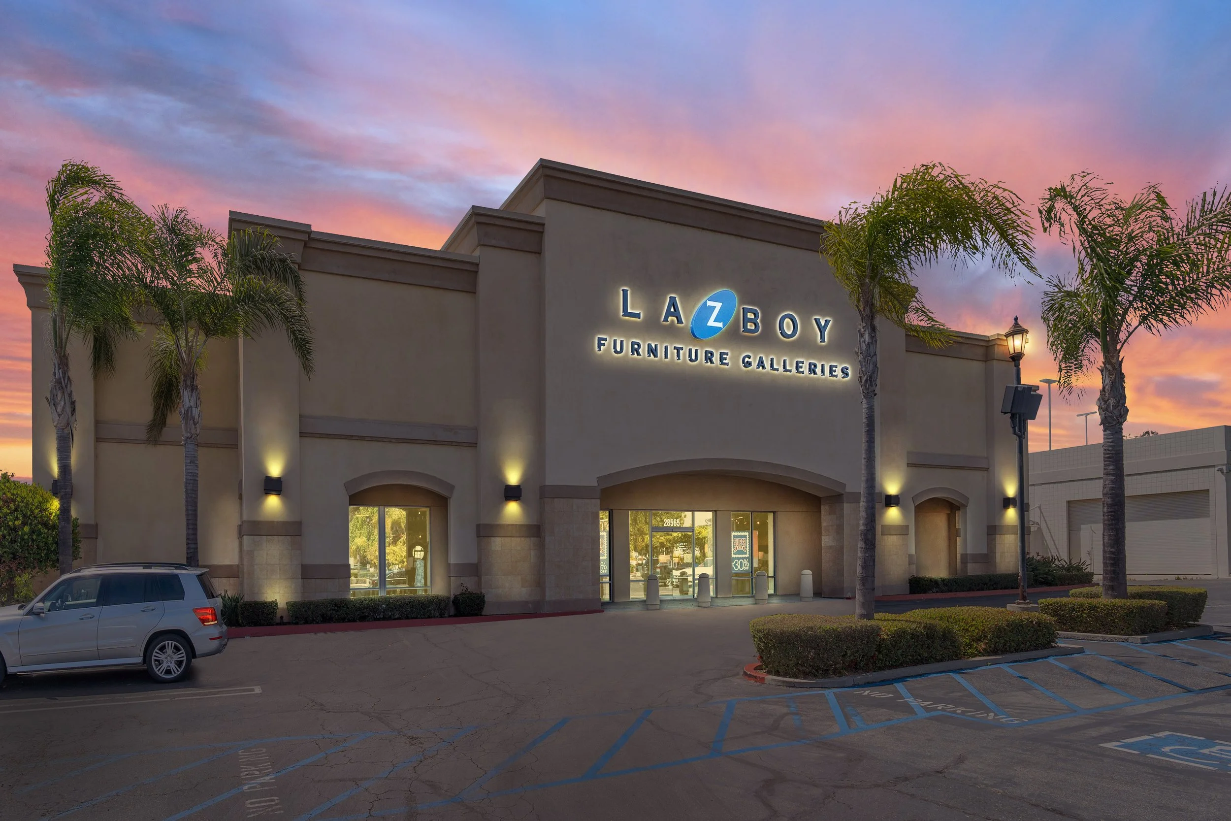 Exterior of Lazboy Furniture Galleries store during sunset with illuminated sign, palm trees, parked car, and marked parking spaces.