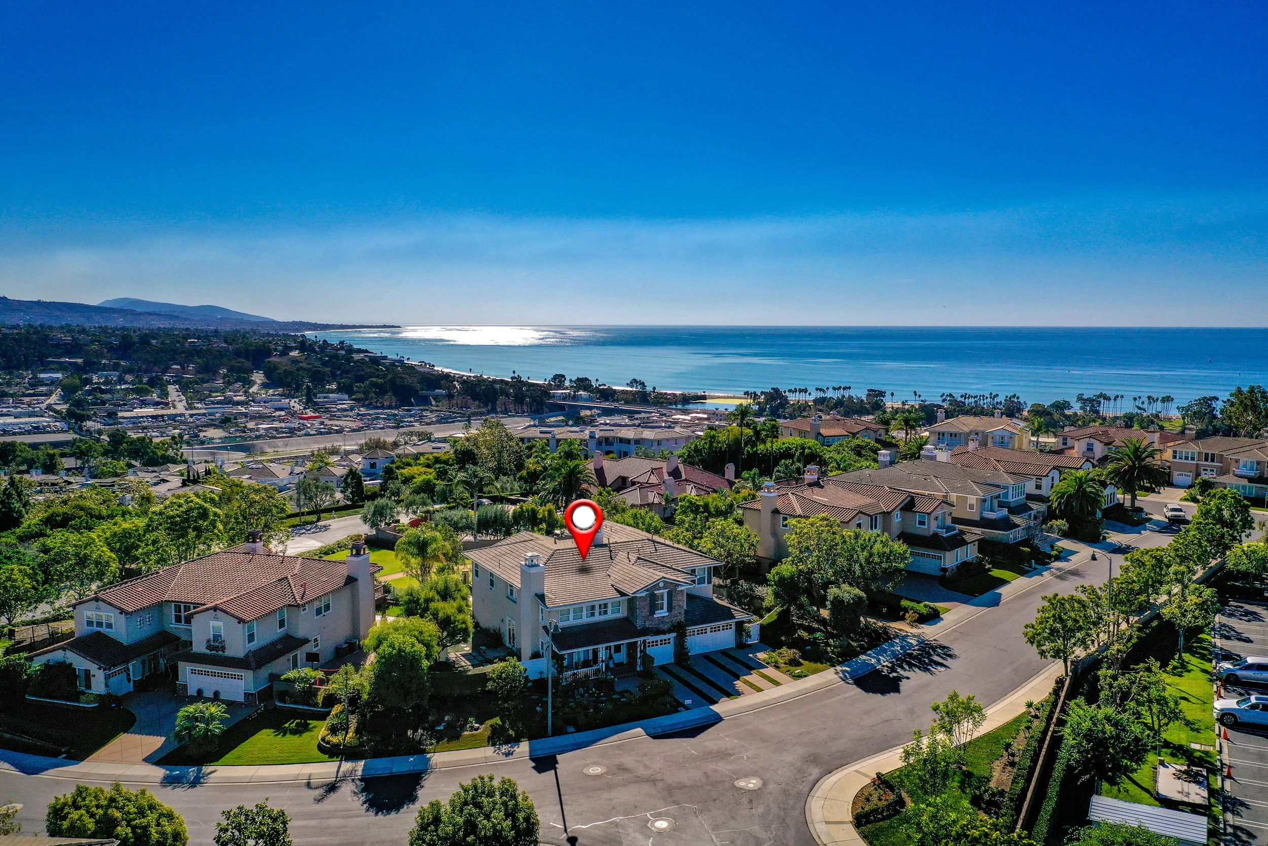 Aerial view of a residential neighborhood with houses, trees, and streets, overlooking a beach and ocean under a clear blue sky.