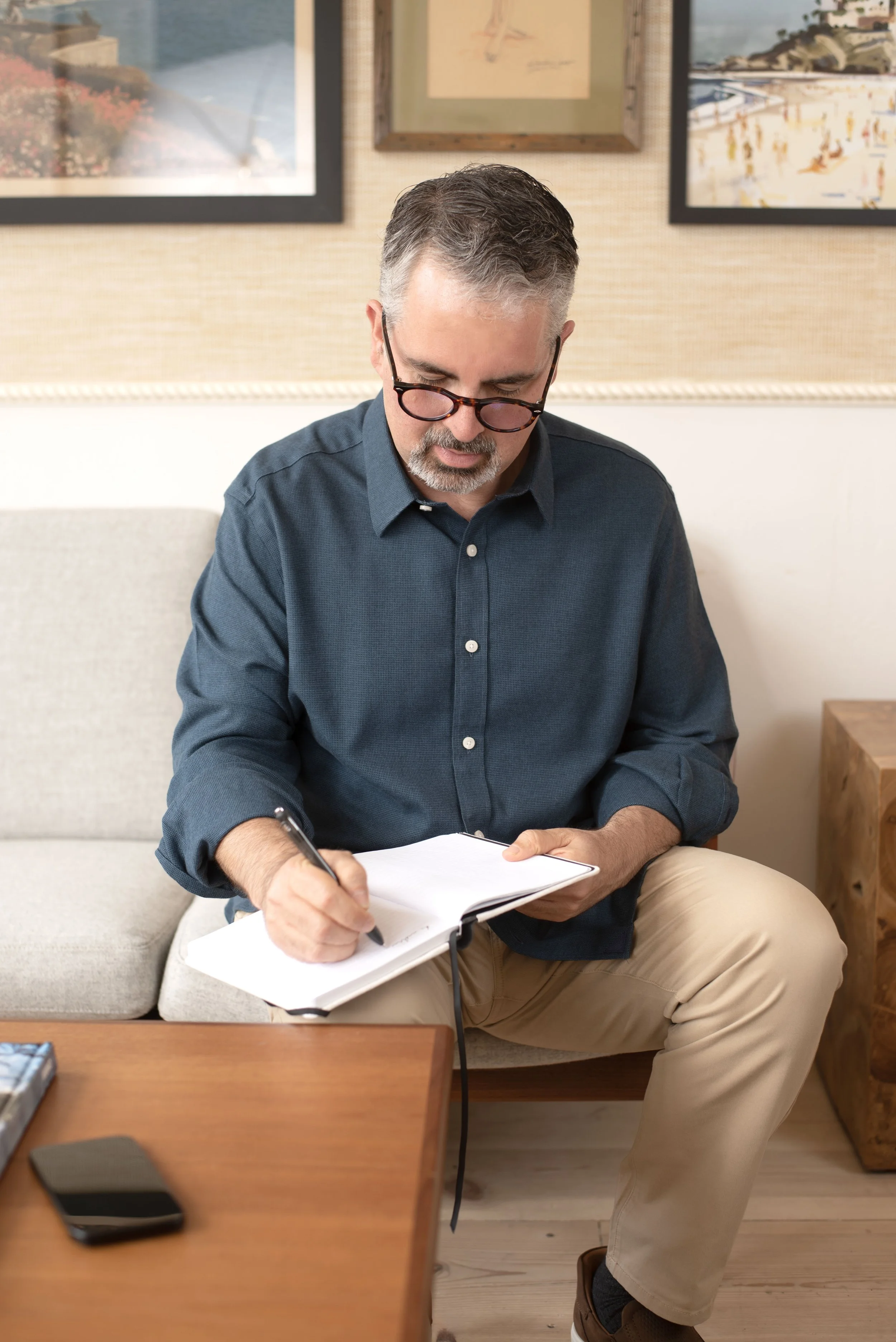 A man with gray hair, glasses, and a beard sitting on a beige sofa writing in a notebook, with a smartphone on a wooden table in front of him and framed artwork on the wall behind him.