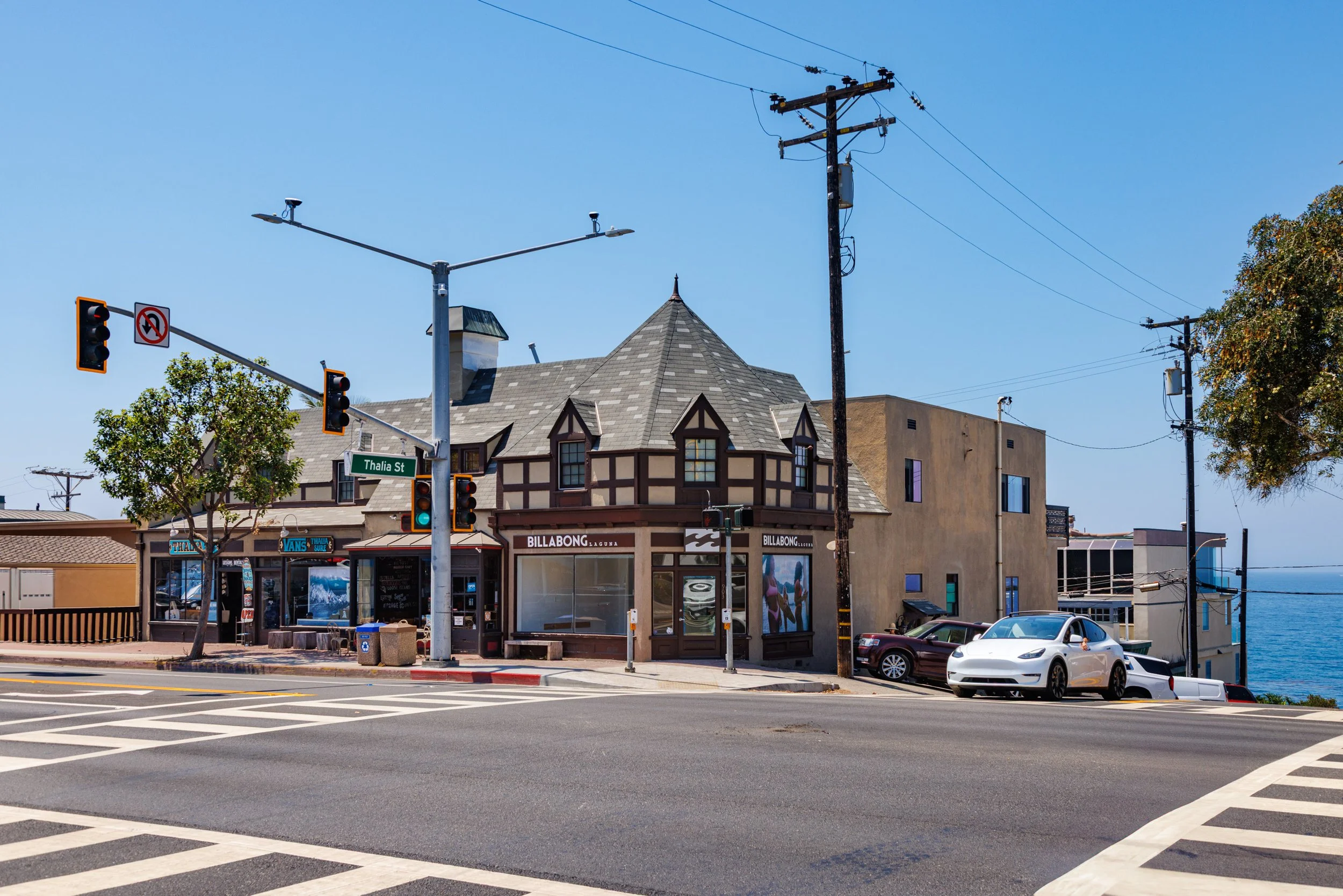 Street corner view with a Tudor-style building housing a Billabong store, traffic lights, street signs, parked cars, and power lines near the ocean under a clear blue sky.