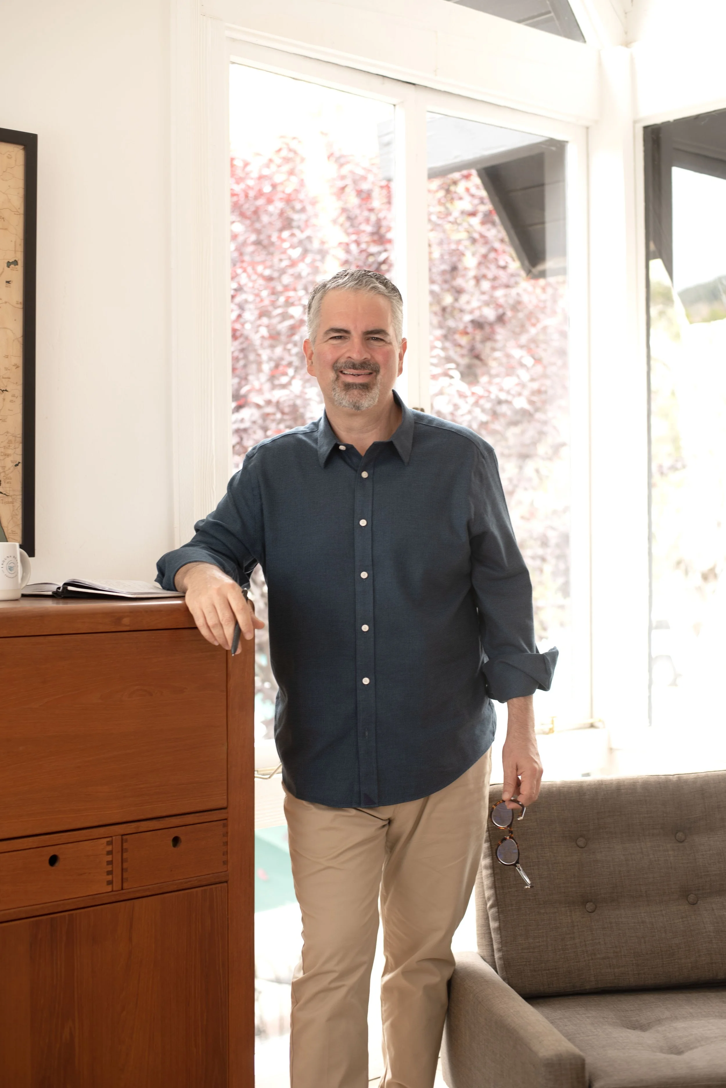 A middle-aged man with gray hair and a beard standing indoors near a wooden cabinet, holding sunglasses, smiling, with large windows and trees visible in the background.
