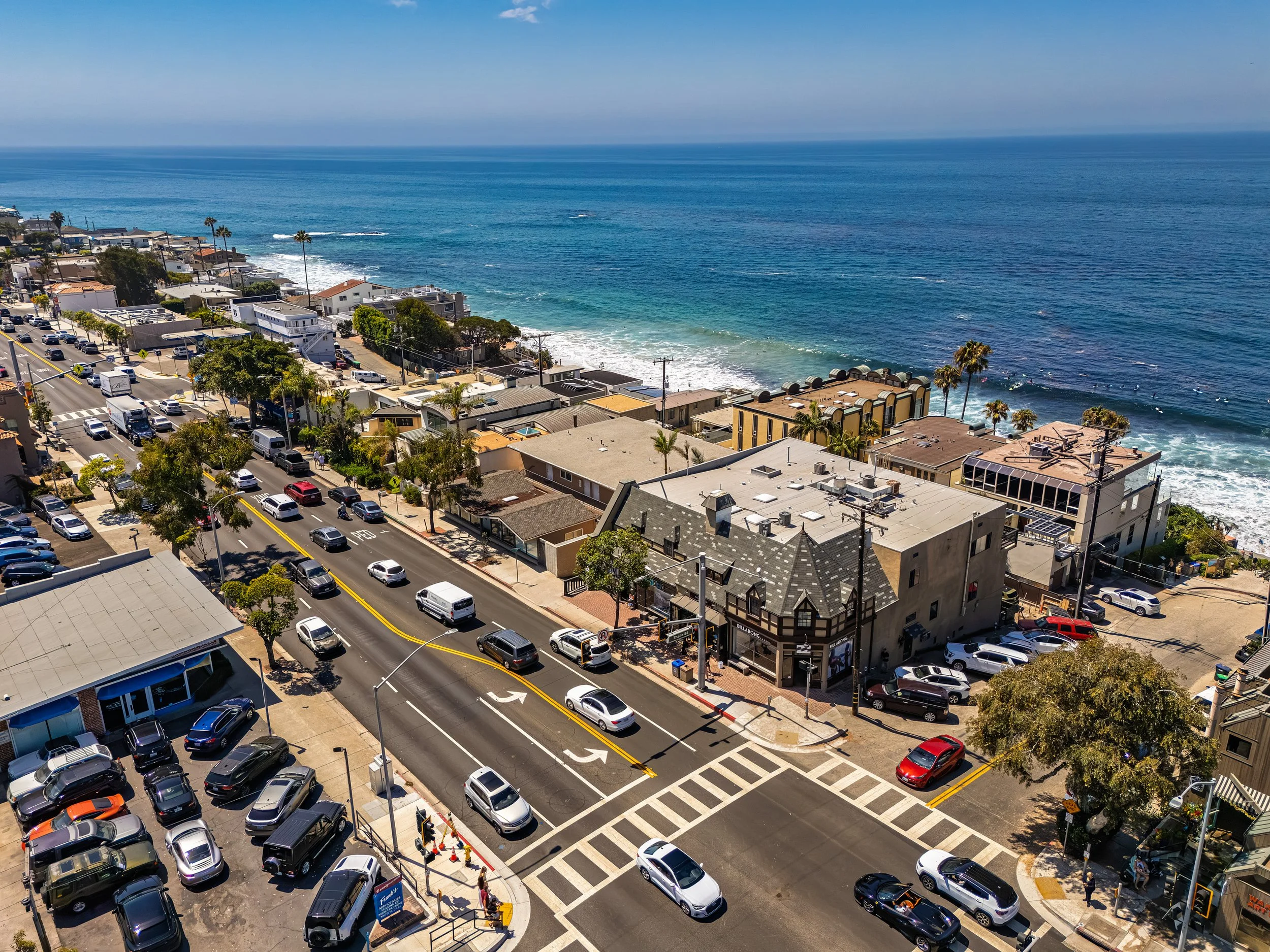 Aerial view of a coastal town with a busy street, parked cars, trees, and buildings near the shoreline with waves and the ocean in the background.