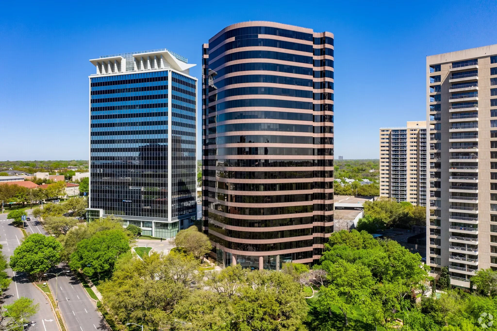 Three modern high-rise office buildings surrounded by trees and streets in an urban area under a clear blue sky.