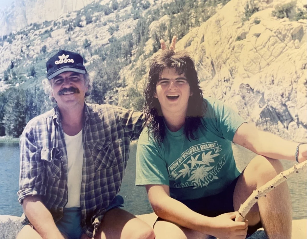 Two people enjoying a sunny day outdoors near a body of water with rocky hills in the background. One is a man wearing a plaid shirt, white T-shirt, black Adidas cap, and smiling. The other is a young woman with long dark hair, wearing a light green T-shirt and smiling. She is holding a stick, and the man is playfully making bunny ears behind her head.