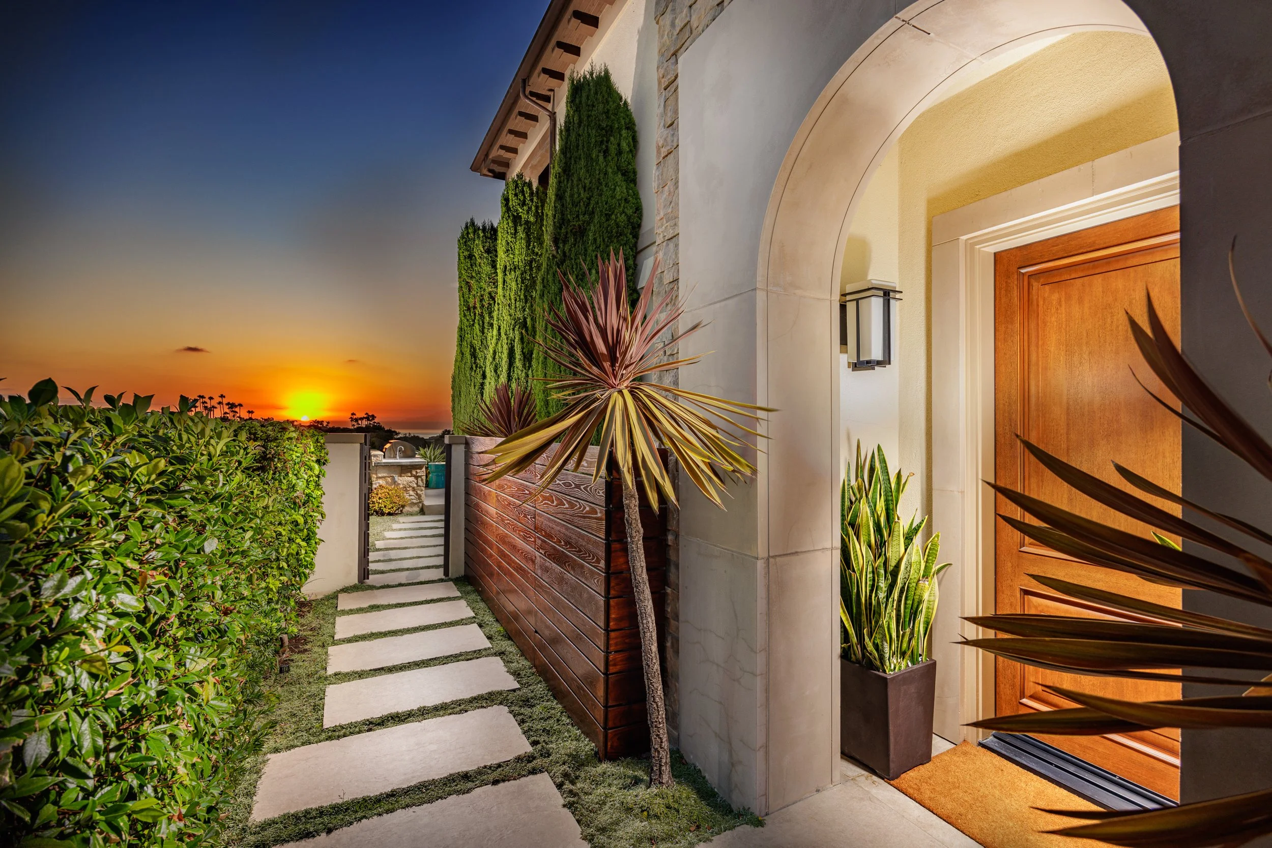 Front entrance of a house with a wooden door, potted plants, stone pathway, lush green shrubs, tall cypress trees, and a sunset sky.