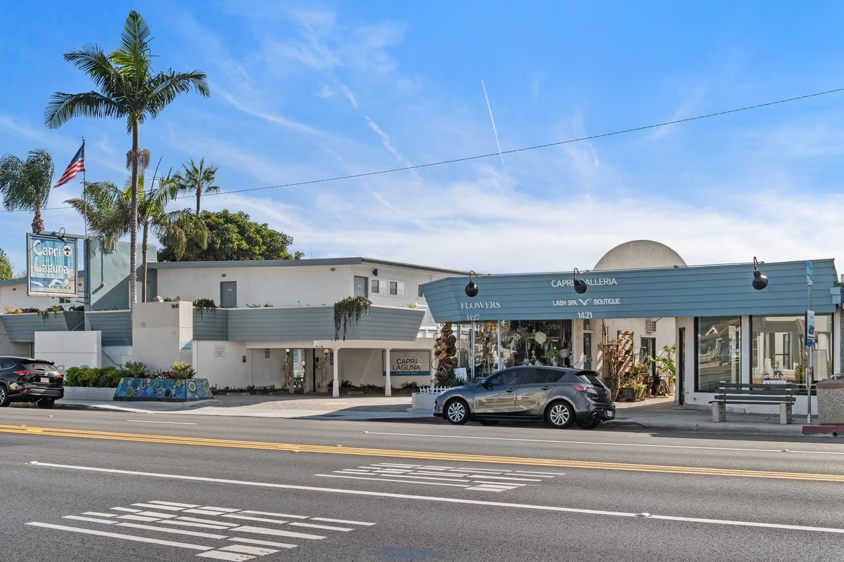 Street view of a shopping area with palm trees, a car parked on the road, and a building with signs for Capri Laguna, flowers, lash spa, and boutique. Clear blue sky with a jet contrail.