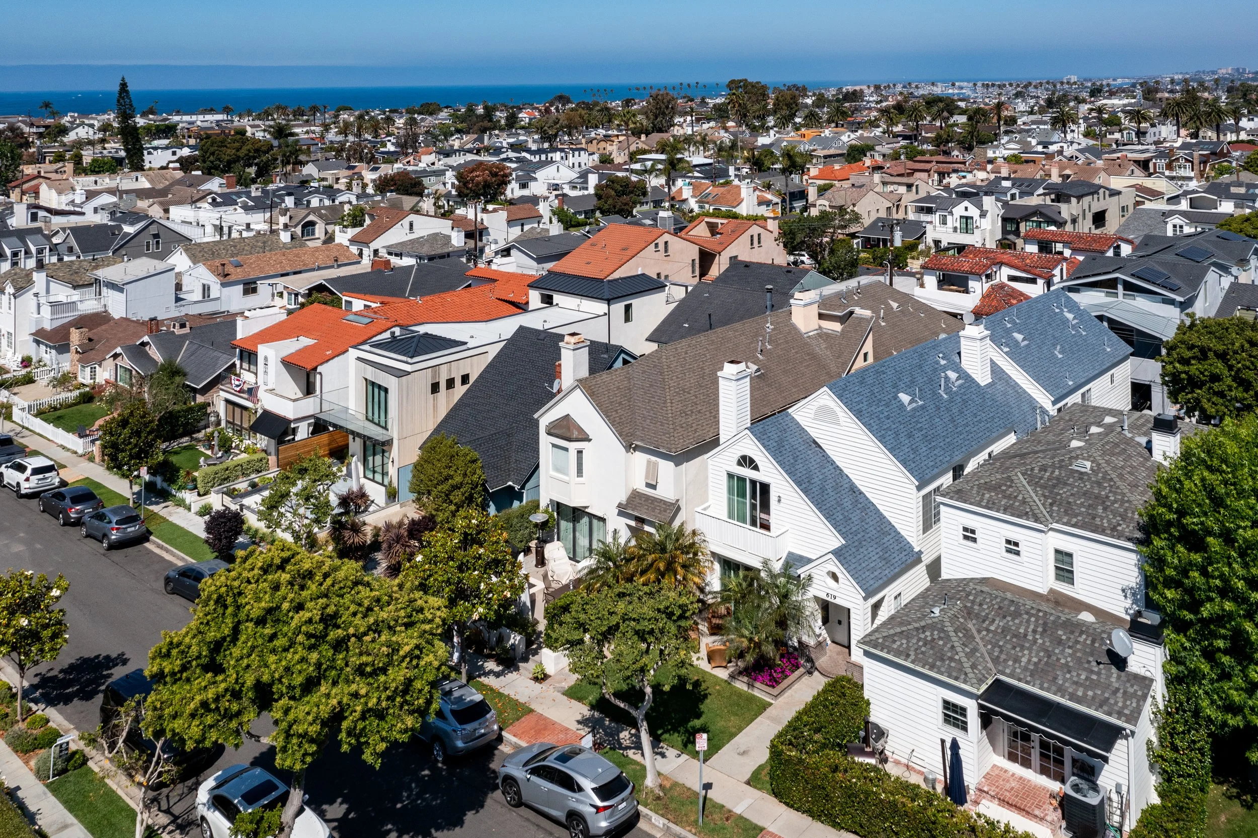 Aerial view of a coastal suburban neighborhood with single-family homes, green trees, parked cars, and the ocean in the background on a sunny day.