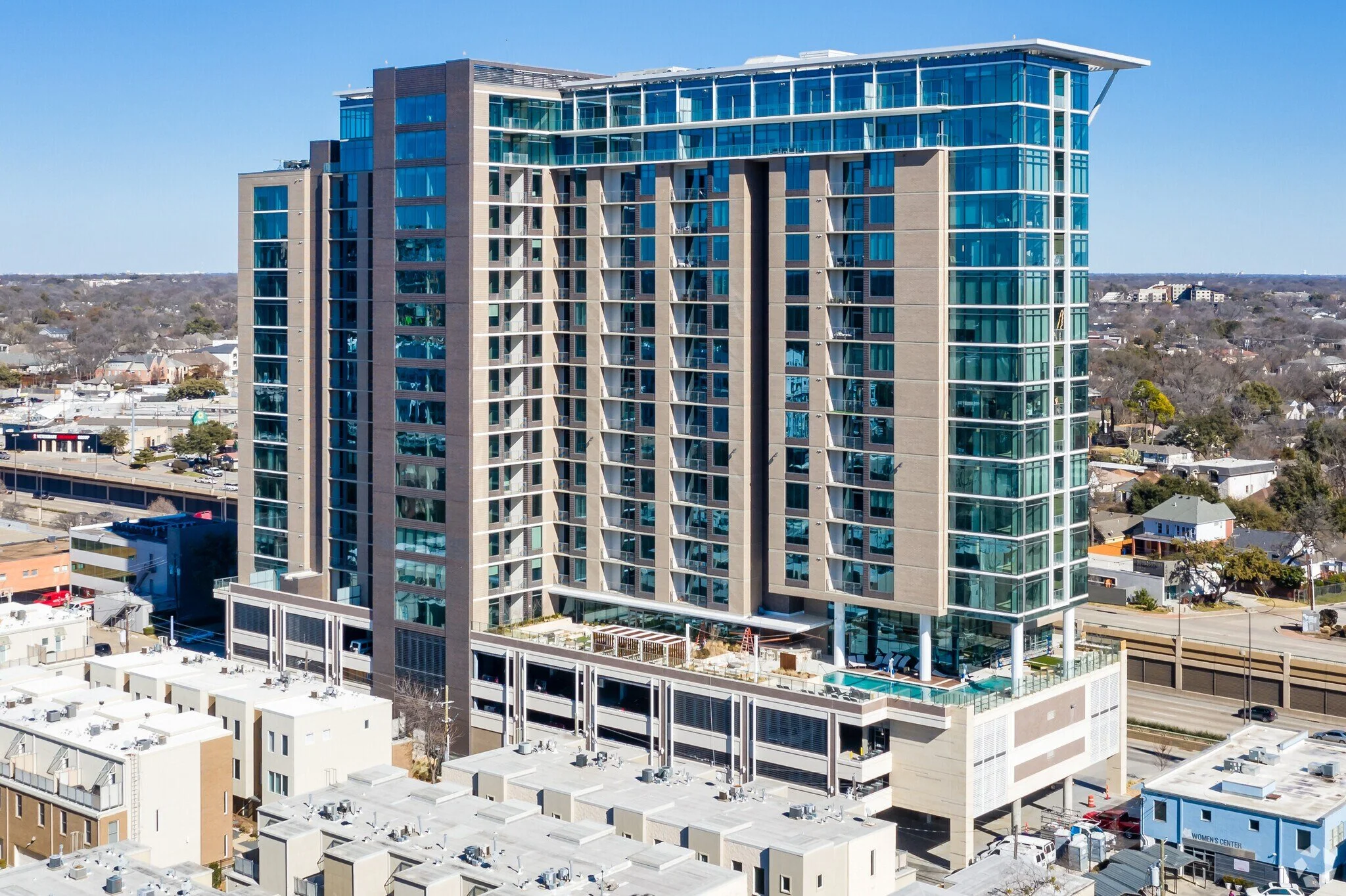 Modern high-rise apartment building with glass windows and balconies, situated in an urban area with smaller commercial and residential buildings nearby.