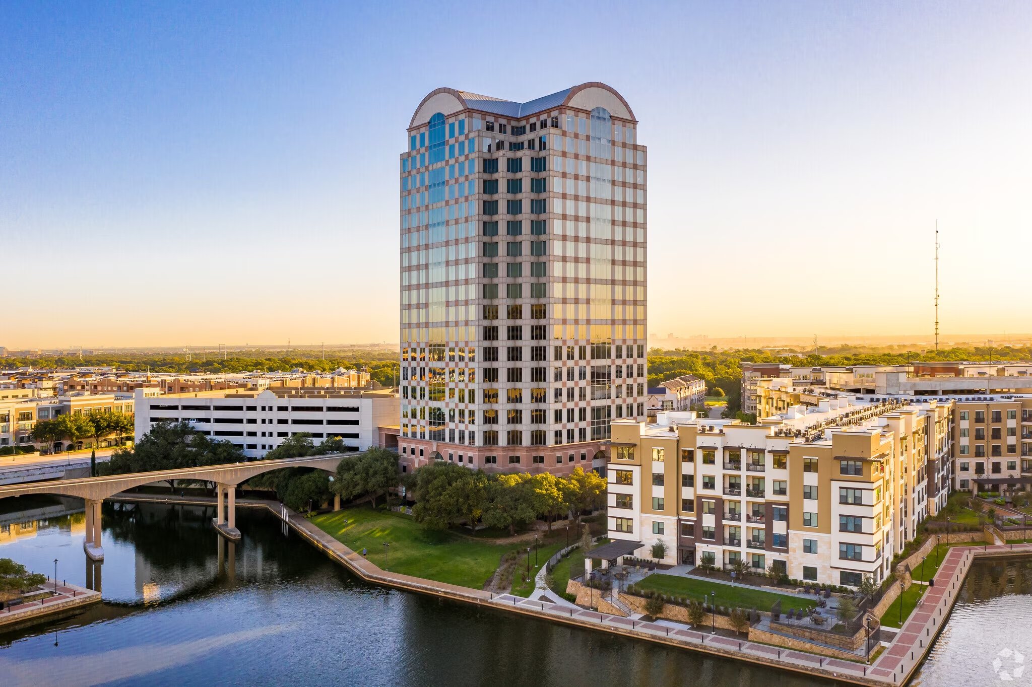 Tall modern high-rise building with reflective glass windows surrounded by smaller buildings, a waterway, and greenery during sunset.