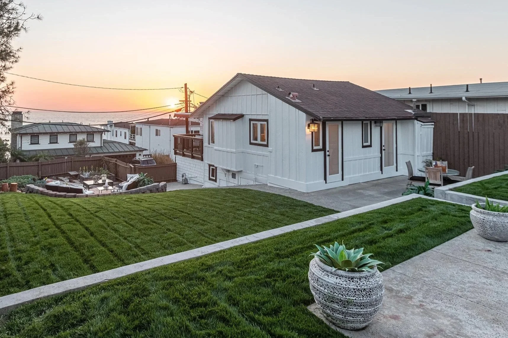 Sunset view of a backyard with a house, outdoor patio, and lawn with potted plants.