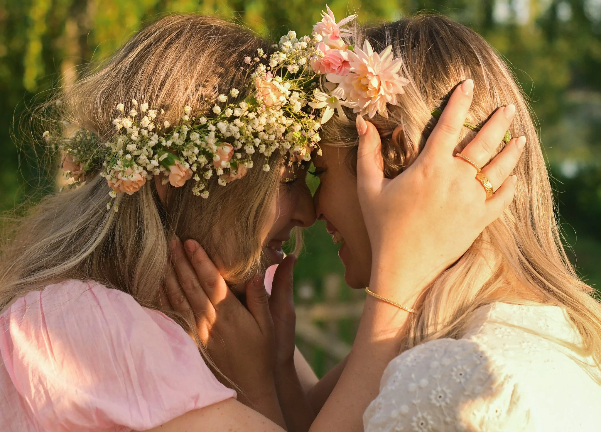 Two women with flowers in their hair touching foreheads and smiling outdoors.