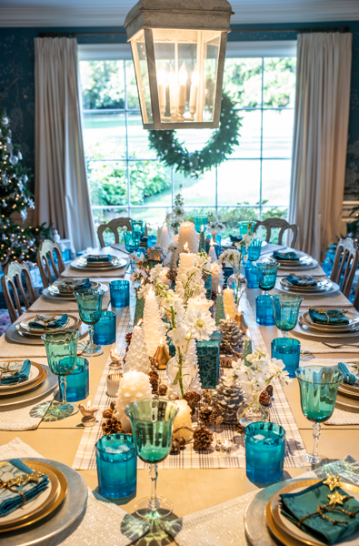 A holiday dining table set with white and gold tableware, blue glasses, and blue napkins, decorated with white flowers and candles, with a festive wreath hanging in front of a window, and a large lantern chandelier above.