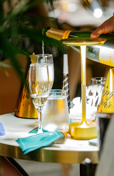 Person pouring champagne into a glass at a New Year's Eve party with hats and decorations.